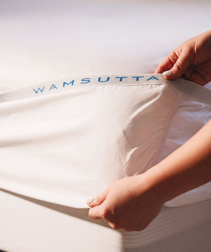 Hands fitting a white sheet onto a mattress, highlighting the branded elastic edge.